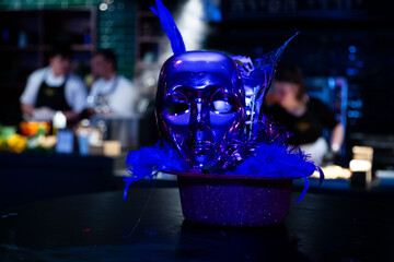 Striking blue feathered mask on glittery surface with blurred chefs in background, invoking a...