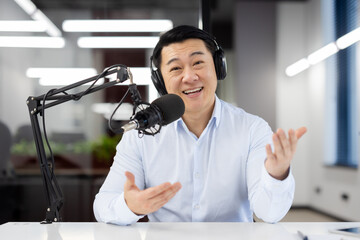 Young Asian man working in office, sitting at desk wearing headphones and in front of desk with microphone and talking to camera via video call. conducts an online meeting, records business training.