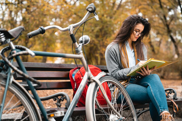Young female student sitting outdoors on the park bench, next to her bicycle and backpack, and...