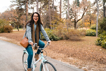 Cheerful young woman with long straight brown hair riding a bike through the city park on her way...