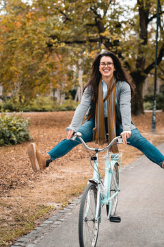 Playful young adult female performing acrobatics while riding her bike in the city park, surrounded by trees in Autumn colors.