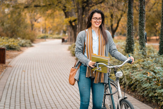 Smiling young entrepreneur holding business papers and pushing her bike while walking in public park during the sunny Autumn day.