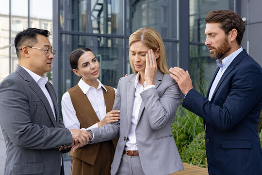 Concerned business colleagues provide comfort to a stressed female professional experiencing a headache outside a modern office building.