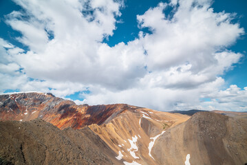 Fototapeta premium Colorful landscape with sunlit pass and sharp rocky mountain of vivid colors under cloudy blue sky. Rocks and sheer crags in sunlight. Snow and shadows of clouds on unusual multicolored mountains.