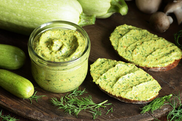 Green zucchini spread in a glass jar with whole wheat bread sandwiches on rustic dark background, healthy summer vegetarian food concept