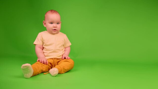A baby dressed in yellow looks around with a curious expression on a green backdrop.