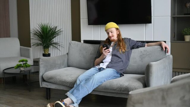A young red-haired guy in a yellow hat sits on the sofa in the living room and scrolls through social networks.