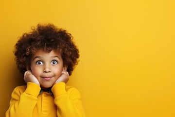 Kid with curly hair wearing a yellow sweater expresses surprise and delight against a yellow background - obrazy, fototapety, plakaty