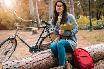 Fototapeta na wymiar Thoughtful young female student with reading glasses studying for exam while sitting on the tree log in natural environment.