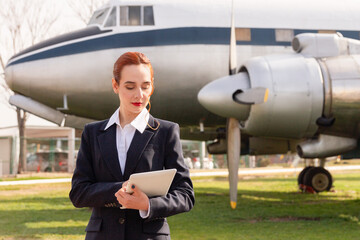 A poised air hostess in a business suit attentively reviews her tablet in front of a vintage airplane, evoking a blend of classic and modern aviation themes