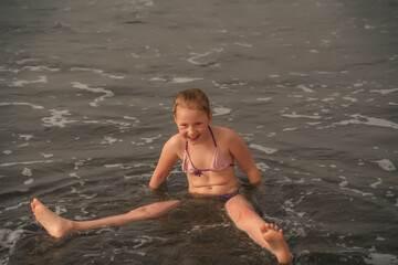 A young girl in a purple bikini laughs as she sits in the shallow seashore waters, waves lapping around. Girl Laughing in Gentle Seashore Waters