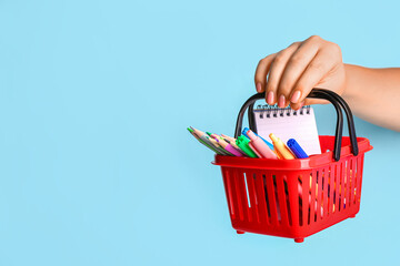 Female hand with red shopping basket and different school stationery on blue background