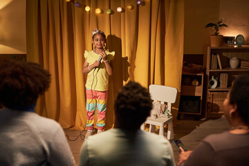 Portrait of Black little girl giving concert or talent show at home in living room with makeshift...