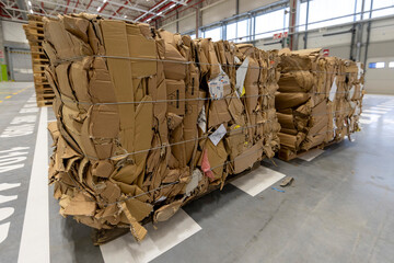 Large bundles of compressed cardboard waste tied with metal wires, ready for recycling, in an industrial warehouse setting.