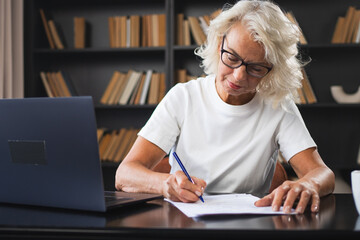 Middle aged senior woman using laptop computer writing notes at workplace. Focused mature old businesswoman work at office making written records doing online work. Mature boss professional worker - obrazy, fototapety, plakaty