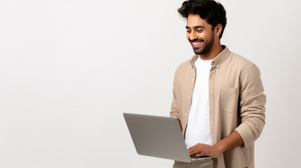 Smiling man holding a laptop, dressed in a beige shirt and a white t-shirt, looking down at his screen, standing against a neutral background.