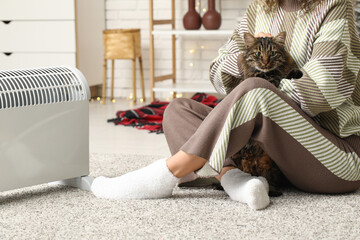 Woman with cute cat sitting on floor near electric heater at home