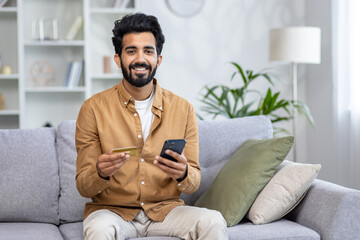 Optimistic diverse male sitting at home on sofa and smiling at camera with cellular phone and...
