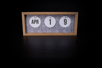 A wooden calendar block showing the date April 19th on a dark black background, save the date or date of an event concept.