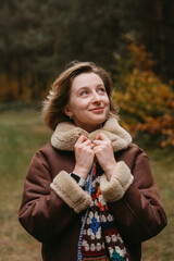 Happy caucasian woman in a brown jacket is standing in the autumn woods and cheerfully looking up