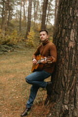 Caucasian adult man plays guitar and leaning on tree in the autumn park