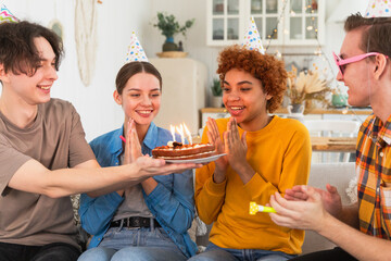 Make a wish. Woman wearing party cap blowing out burning candles on birthday cake. Happy Birthday party. Group of friends wishes girl happy birthday. People celebrating birthday with party at home