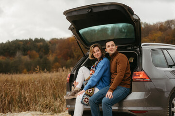 Happy young woman and man sitting in the open trunk of a car while traveling in autumn and looking away, road trip concept