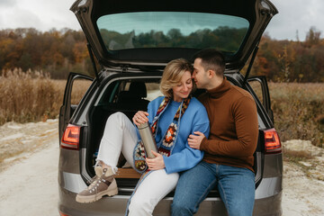 Happy adult couple sitting in the open trunk of a car while traveling in autumn, road trip concept, man embracing woman
