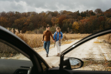 Happy man and woman are walking down a country road next to their car holding hands, adult couple on road trip in autumn