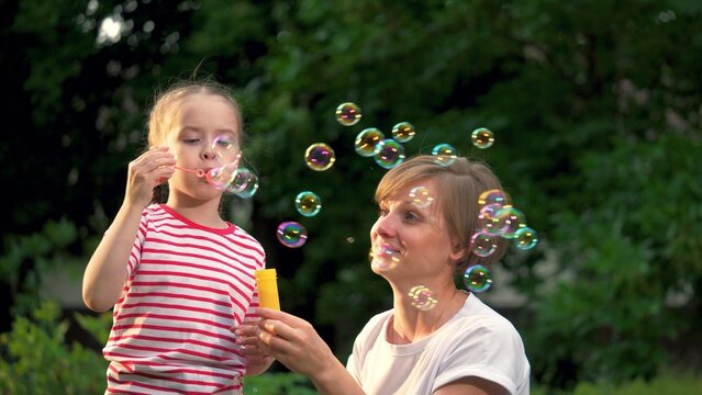 Little girl and mother blow soap bubbles together in green city park. Mother with joyful little girl plays soap bubbles walking in park. Little girl and mother rest blowing soap bubbles in garden