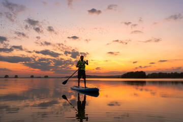 silhouette of man standing on inflatable SUP board and paddling through shining water surface. Wide...