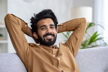 Positive muslim guy leaning on melange couch with hands behind head and smiling at camera on...