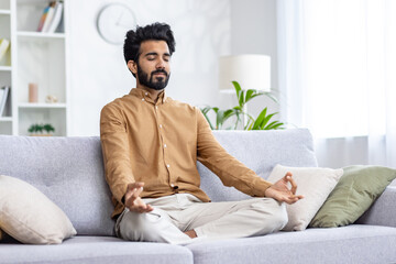 Calm middle eastern man sitting on pillowed sofa in siddhasana position and using hands mudra with...