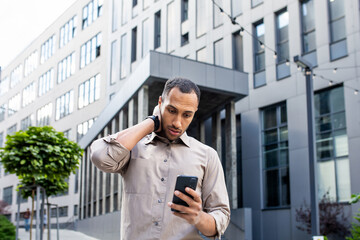 Disturbed arab man rubbing head with hand while looking at mobile phone screen on background of office building. Stressed manager leaving corporate workplace and receiving text message from boss.