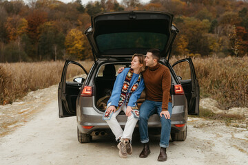 Happy young woman and man sitting in the open trunk of their car while traveling in autumn and looking away, couple road trip concept