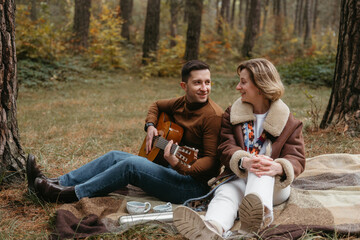 A cheerful man is playing a guitar for a happy woman while they are sitting on a blanket in the autumn woods