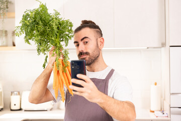 long-haired man cooking at home looking at tablet, chopping vegetables, taking selfie photos, carrots in his hand