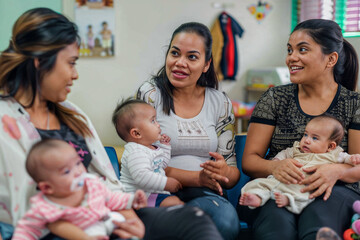 Diverse group of women with their babies in a pediatrician's office waiting area, engaging in conversations and sharing laughter.

