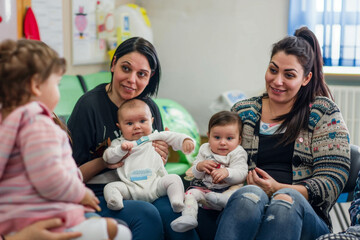 Diverse group of women with their babies in a pediatrician's office waiting area, engaging in conversations and sharing laughter.

