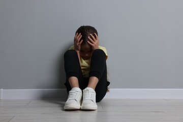 Child abuse. Upset girl sitting on floor near grey wall
