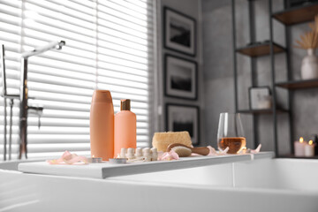 Wooden tray with wine, toiletries and flower petals on bathtub in bathroom