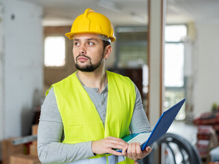 Portrait of a qualified builder on a construction site indoors, standing, holding a laptop