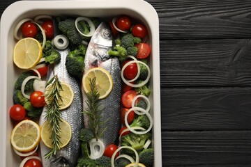 Raw fish with vegetables and lemon in baking dish on black wooden table, top view. Space for text
