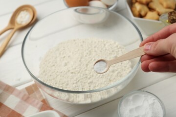 Woman taking spoon with baking powder from bowl at white wooden table, closeup
