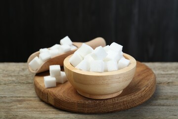 Fototapeta na wymiar White sugar cubes in bowl and scoop on wooden table against dark background, closeup