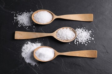Natural salt and wooden spoons on black table, flat lay