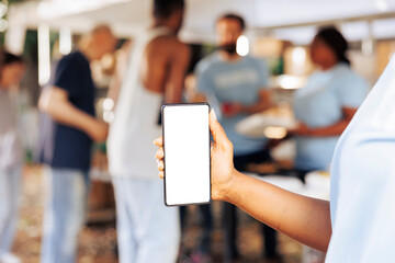 Close-up shot of african american person holding a mobile device showing blank copyspace mockup template. Detailed image of a smartphone with isolated white screen display at non-profit food drive. - obrazy, fototapety, plakaty