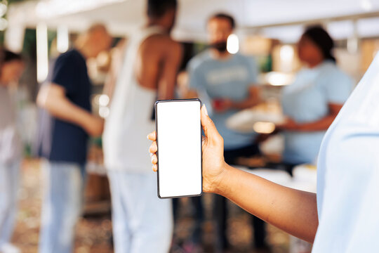 Close-up shot of african american person holding a mobile device showing blank copyspace mockup template. Detailed image of a smartphone with isolated white screen display at non-profit food drive.
