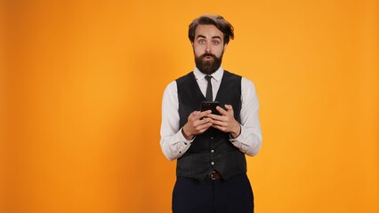 Diner employee messagging on smartphone, posing against yellow background. Experienced butler in...