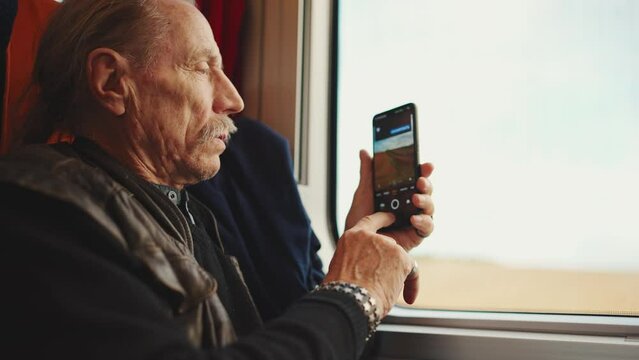 An old man takes a photo of the view from a train window using a smartphone while traveling. Senior rides on a train and films nature on a mobile phone, Slow motion, Morning light